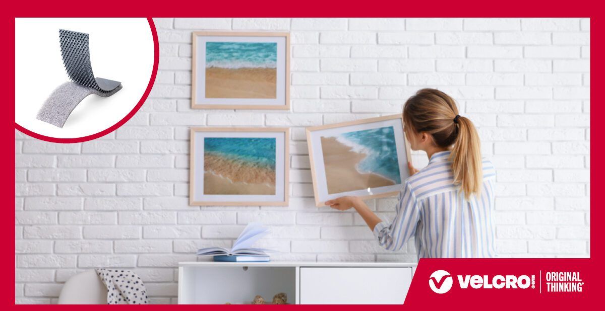 A woman in a striped shirt hangs a framed beach photo on a white brick wall using VELCRO® Brand fasteners. Two other framed beach prints are already mounted. An inset shows hook and loop strips strips. The 'VELCRO® Brand ORIGINAL THINKING™' appear in the corner.