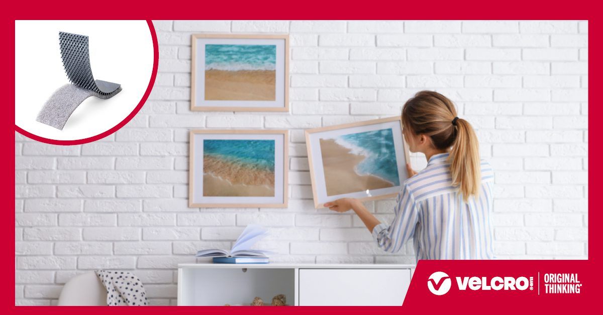 A woman in a striped shirt hangs a framed beach photo on a white brick wall using VELCRO® Brand fasteners. Two other framed beach prints are already mounted. An inset shows hook and loop strips strips. The 'VELCRO® Brand ORIGINAL THINKING™' appear in the corner.