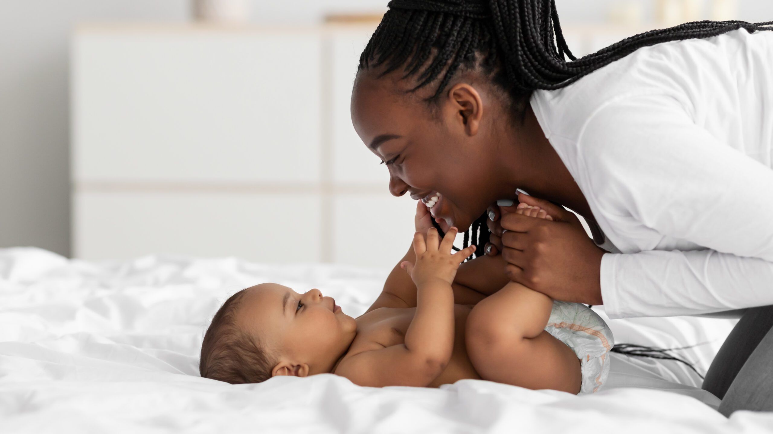 Mom and baby in a diaper smiling and playing together on a bed.